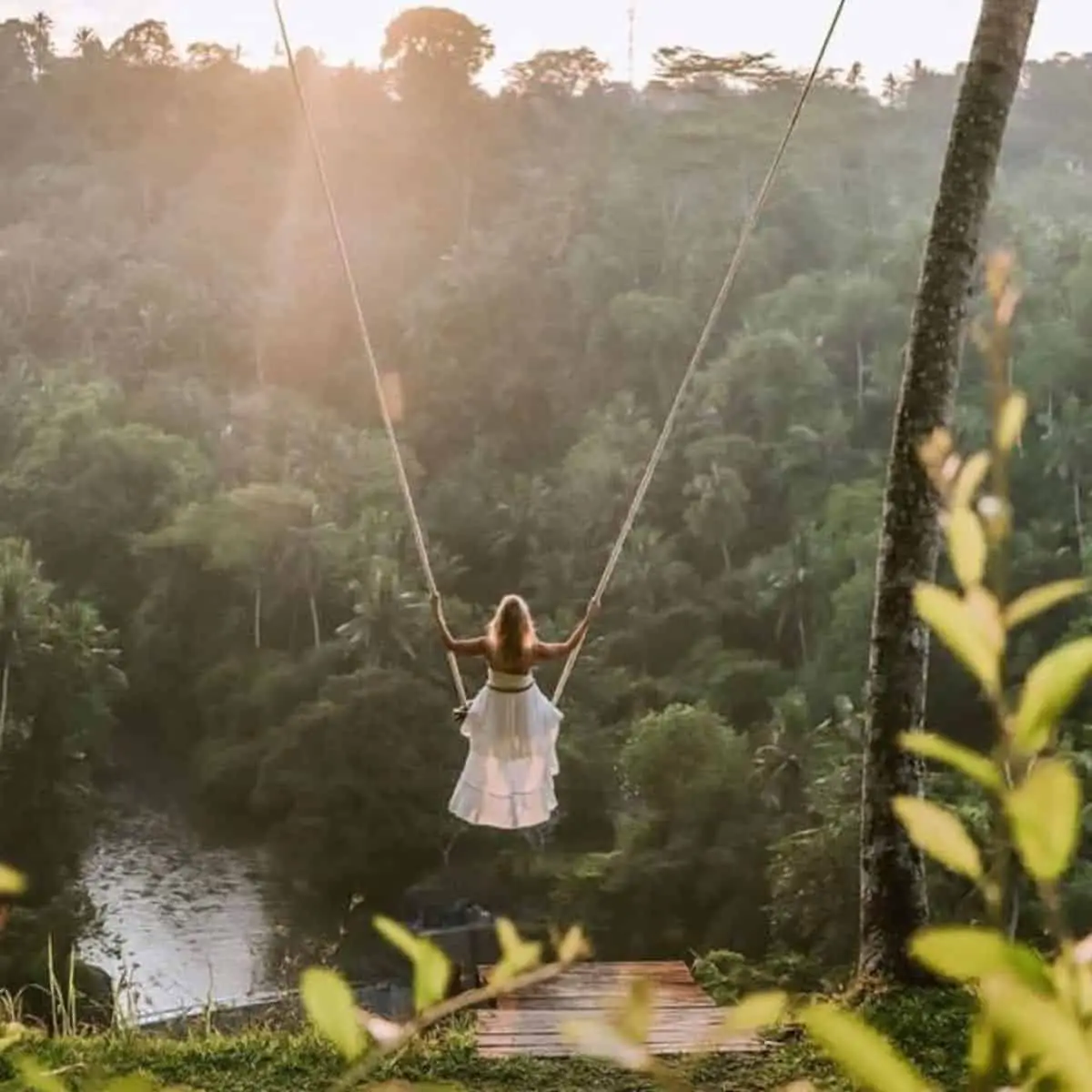 A woman in white dress riding in Zen Hideaway’s swing without harness with a forest and river view in front