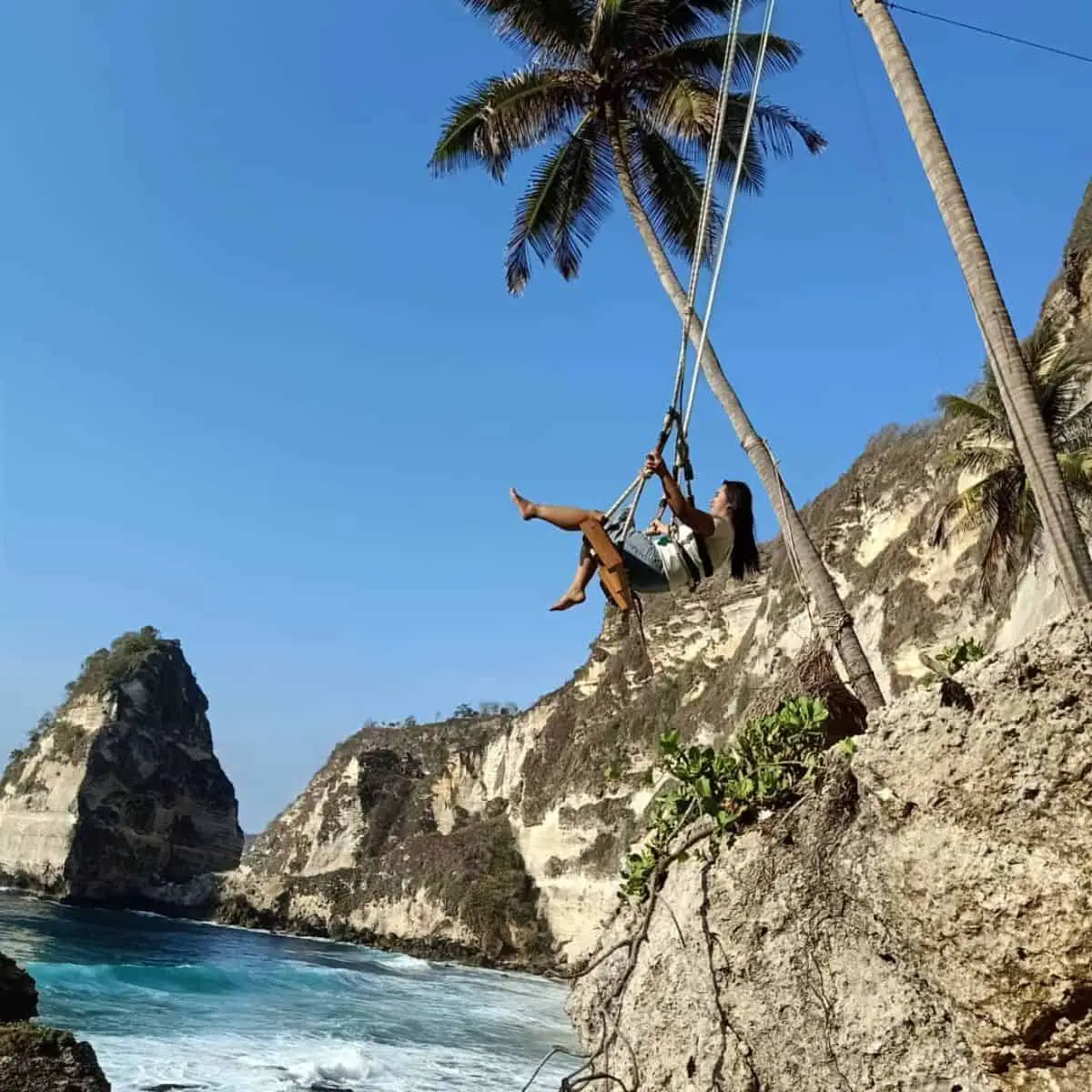 A woman swinging in Nusa Penida with blue waters underneath and rock formations surrounding the beach
