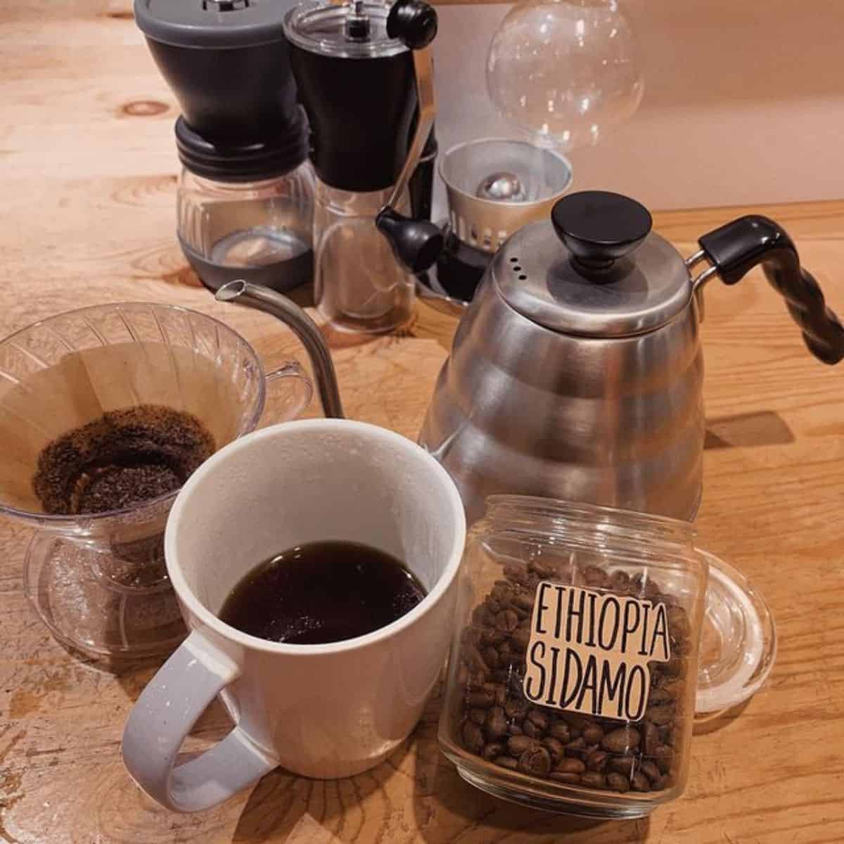 https://tabitogether.com/wp-content/uploads/2023/02/Coffee-beans-in-a-transparent-jar-with-a-cup-of-coffee-and-other-coffee-maker-equipments-laid-in-the-table-of-PBC-Coffee.jpg