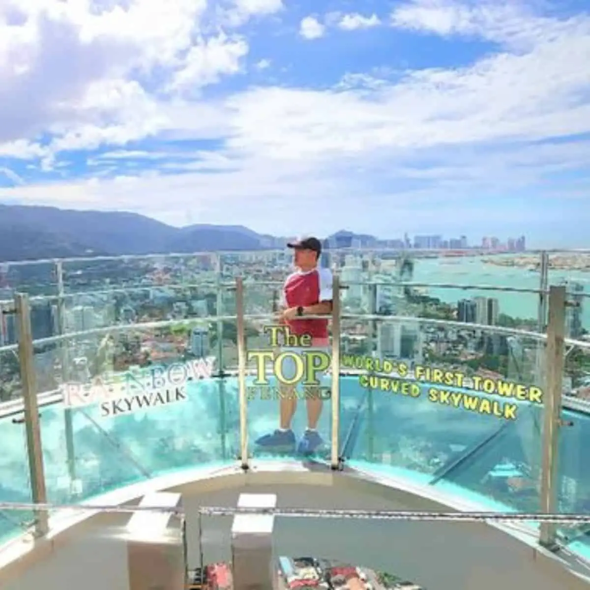 A guy standing at The Top’s Rainbow Skywalk with glass floor and tall buildings below the platform