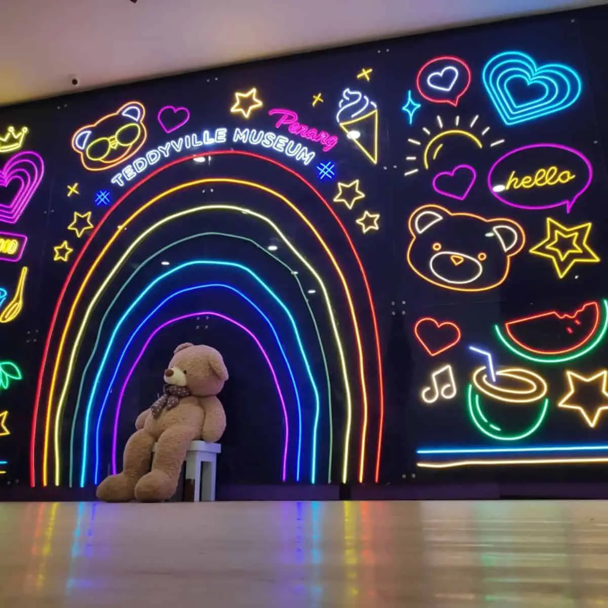 A human size Teddyville Museum’s teddy bear sitting in a chair with colourful lights as a backdrop